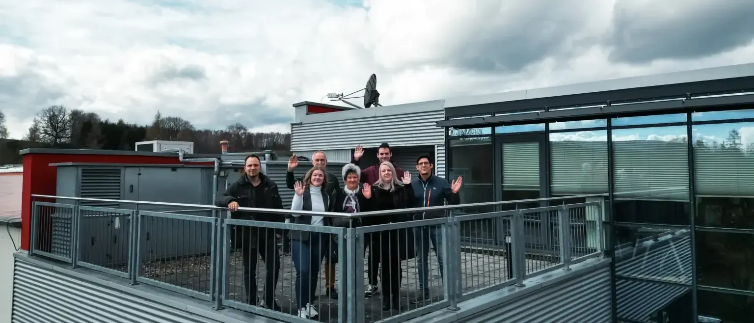 Unsere Kollegen aus der Verwaltung stehen an einer Aussenecke unserer Dachterrasse und schauen zum Betrachter. Vor ihnen das Geländer, im Hintergrund sieht man die verglaste Seite des Gebudes, ein Stück der Fassade der Werkhalle und weiter im Hintergrund eine Baumreihe. Der Himmel ist bewölkt