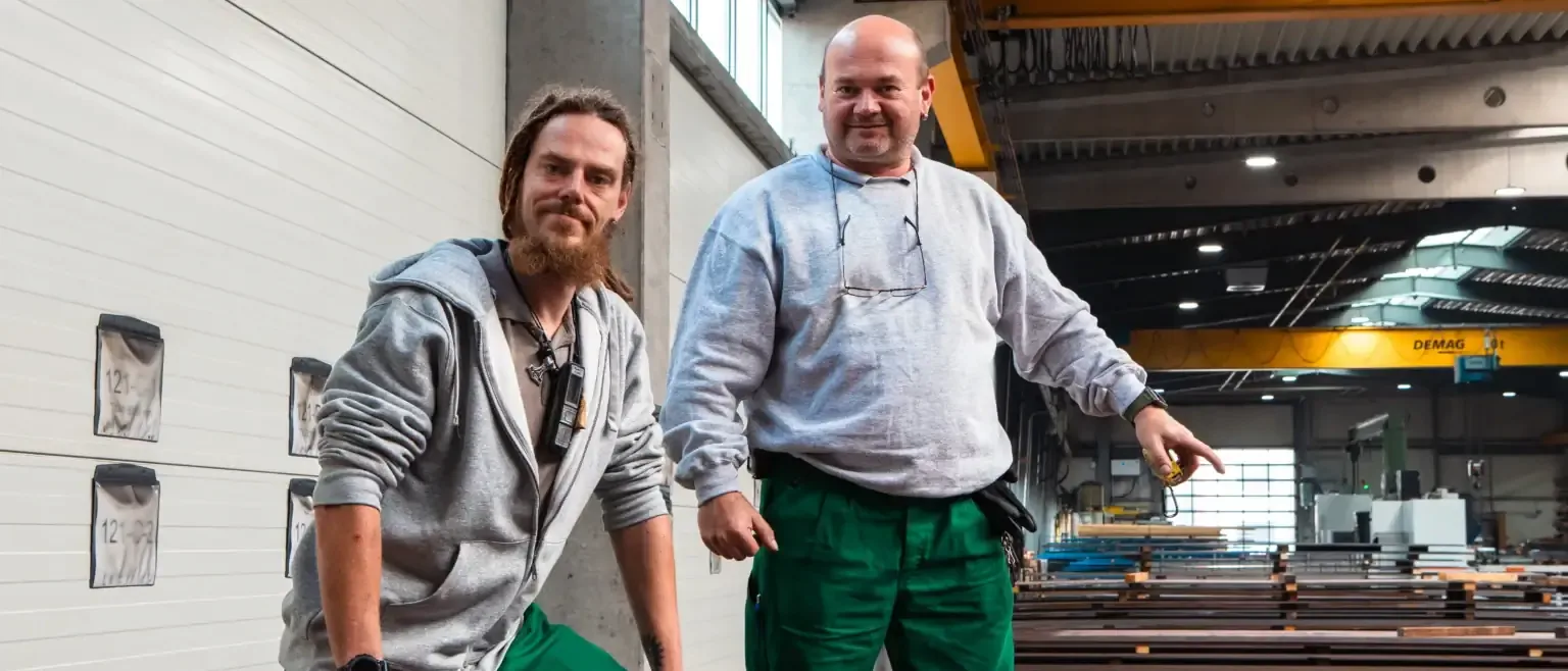 Zwei Kollegen in unserer Produktionshalle, schauen zum Betrachter. Einer deutet stolz auf die Stahlbleche im Hintergrund. Im Hintergrund sind Stahlbleche und ein Portalkran zu sehen