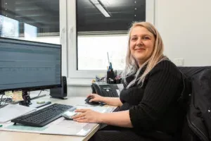 Unsere Ausbilderin in der Verwaltung an Ihrem hellen Arbeitsplatz. Sie sitzt an ihrem Schreibtisch, vor Monitoren und Tastatur. Im Hintergrund sieht man ein riesiges Fenster, welches über eine Außenjalousie verfügt, ein modernes Telefon und Schreibutensilien. 