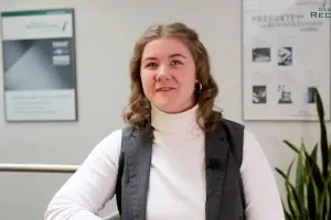 Eine Auszubildende in der Verwaltung, bekleidet mit hellem Pullover und Weste, lehnt an einem Tisch und ist im Gespräch mit einer weiteren Person, die nicht im Bild zu sehen ist. Im Hintergrund ist eine weiß tapezierte Wand, mit eineigen  Bilderrahmen, die Informationen über die Firmengeschichte zeigen.