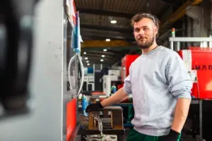 Einer, unserer Auszubildenden in unserer Produktionshalle steht an einer Maschine, die er bedient. Im Hintergrund sieht man weitere Maschinen und einen Portalkran