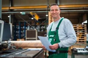 Ein Versandmitarbeiter schaut lächelnd zum Betrachter. Er steht vor einer Werkbank, mit Papieren in der Hand. im Hintergrund sieht man eine Kartonverpackung, und dahinter Einrichtung sowie Säule und Unterzug der Produktionshalle.