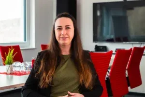 Eine Verkaufsmitarbeiterin sitzt in einem unserer Besprechungsräume, schaut zum Betrachter. Im Hintergrund sind moderne, rote Stühle, ein sehr großer Präsentationsbildschirm, Tisch, Telefon und Fenster, mit dunklem Vorhand zu sehen.